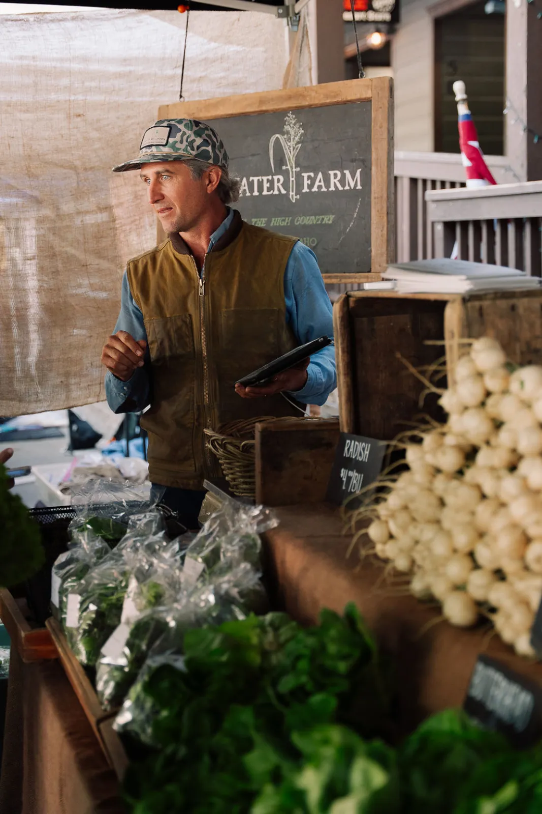 A farm vendor standing among crates of fresh produce and greens